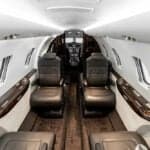 A luxurious private jet interior with dark leather seats, wood flooring, and a view towards the cockpit.