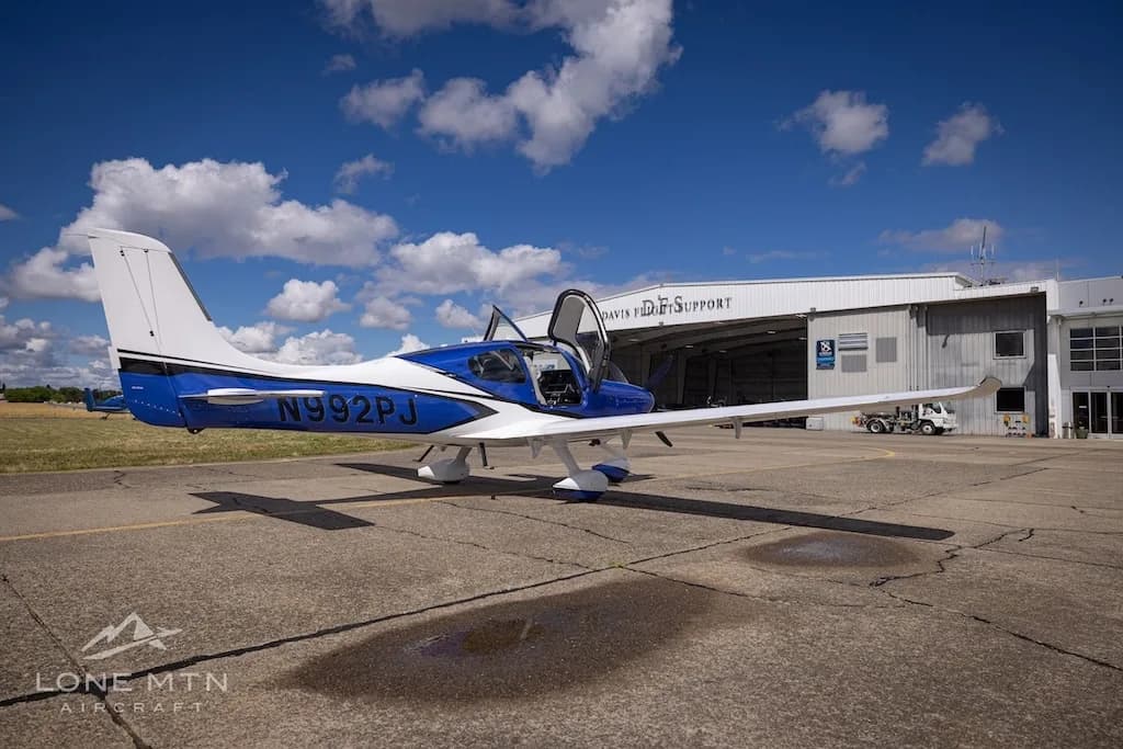 A blue and white Cirrus SR22 aircraft with its door open, parked on an airfield in front of a hangar.