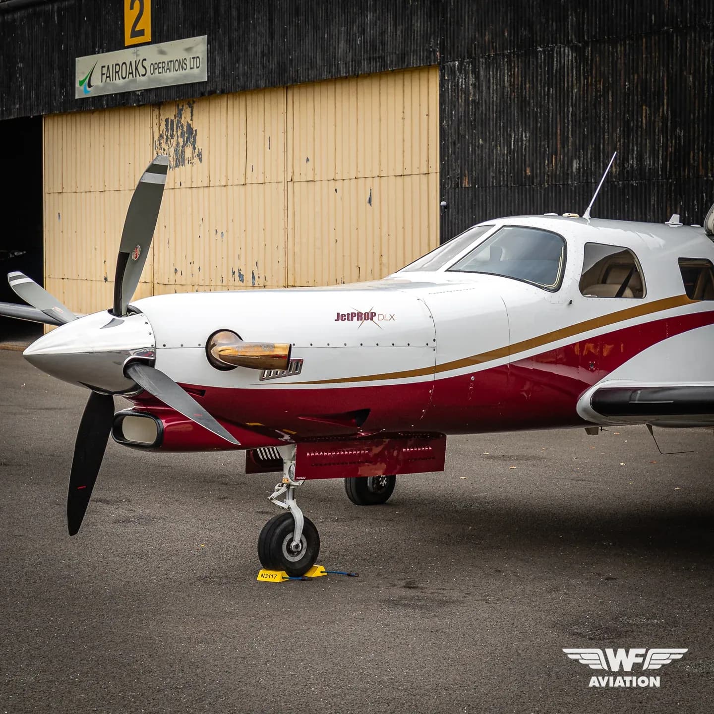 A white JetPROP DLX turboprop aircraft with red and gold stripes, registration N3117, parked on an tarmac in front of a hangar.