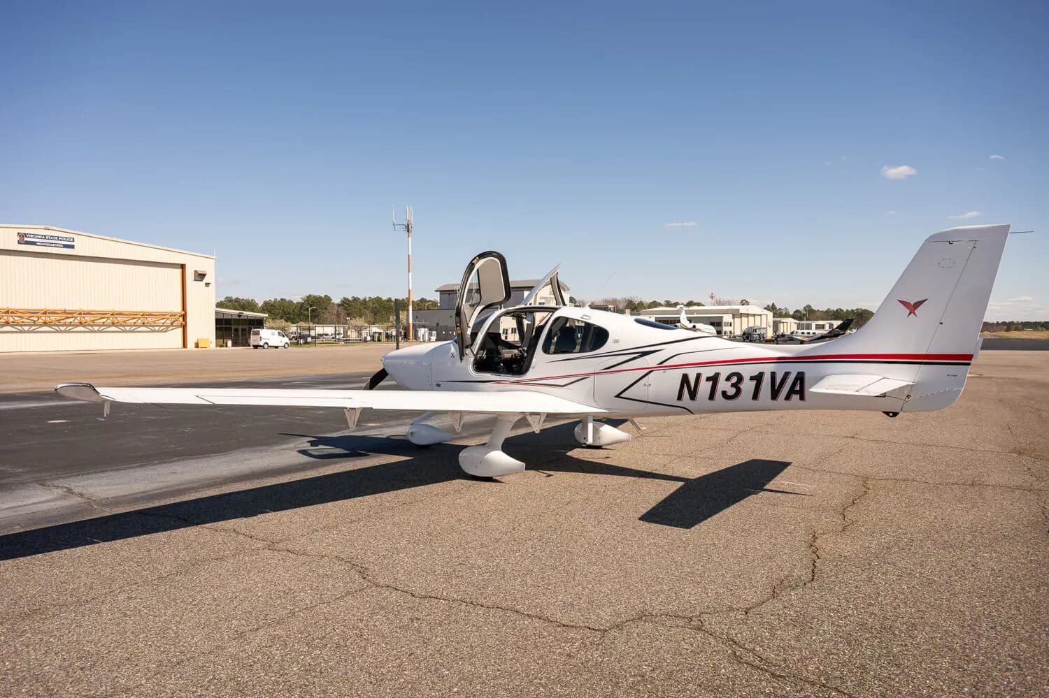 A white Cirrus SR22 aircraft with red and black accent stripes, tail number N131VA, parked on an airfield with both doors open and hangars in the background.
