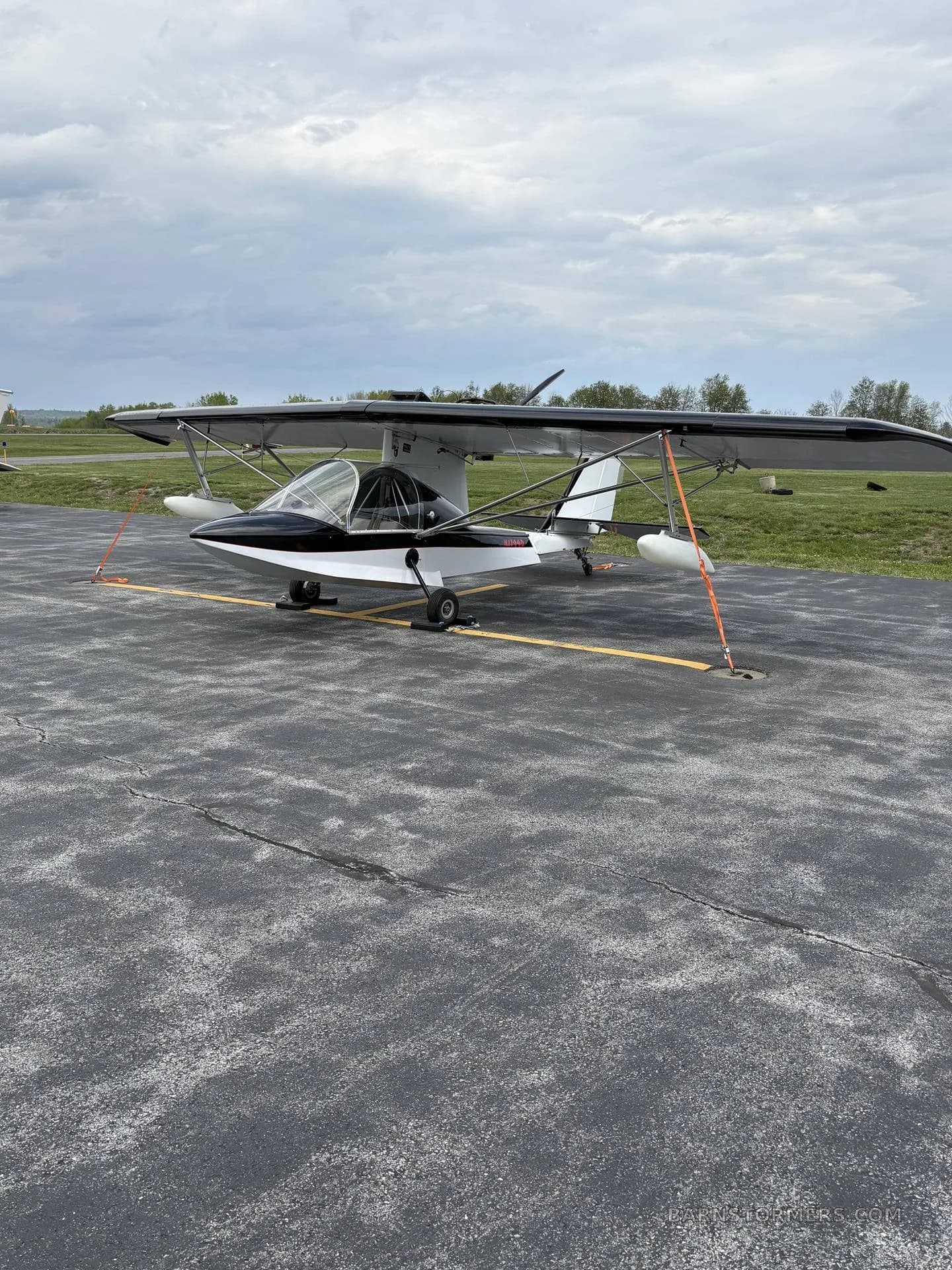 A white and black ultralight aircraft with registration N7749D tied down on an asphalt apron under a cloudy sky.