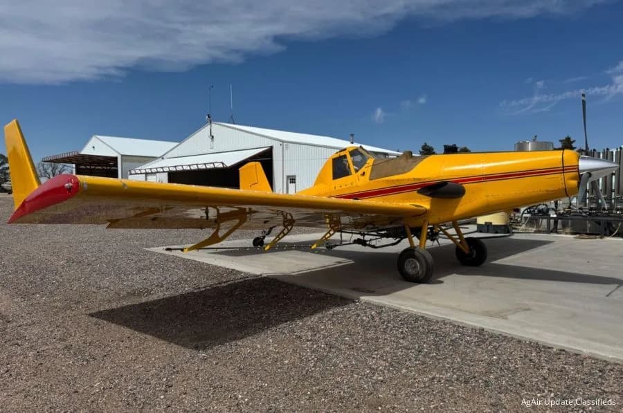 A yellow agricultural aircraft with red stripes and a red tail, parked on a concrete pad next to a hangar under a blue sky.