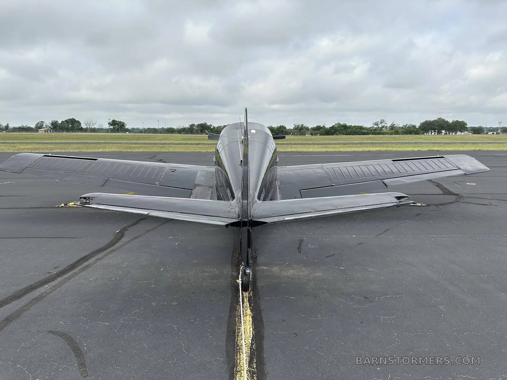 A rear view of a black single-engine aircraft with a V-tail, parked on an asphalt runway under a cloudy sky.
