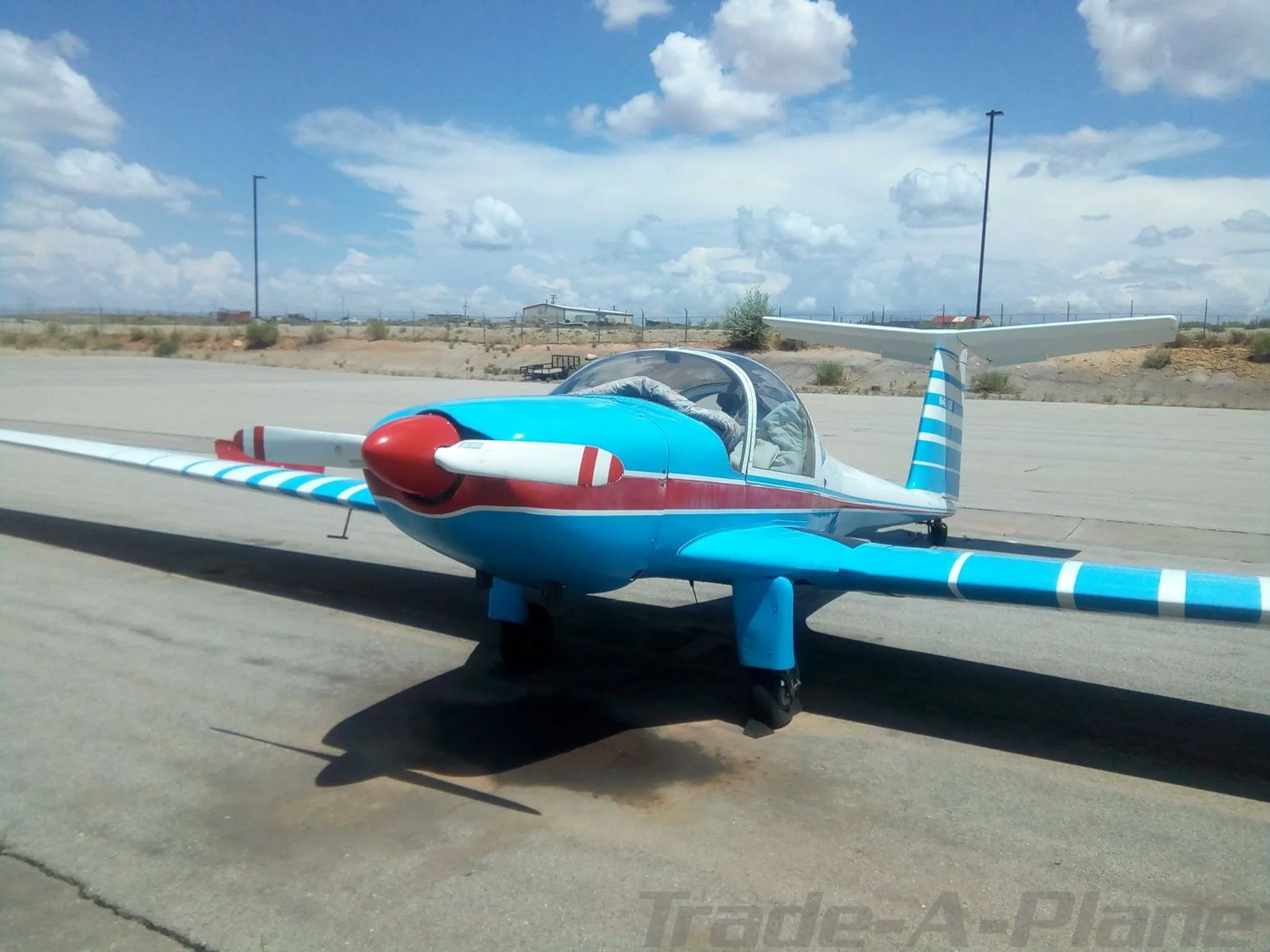 A front view of a blue and white aircraft with a red nose and stripes, parked on an airfield under a partly cloudy sky.