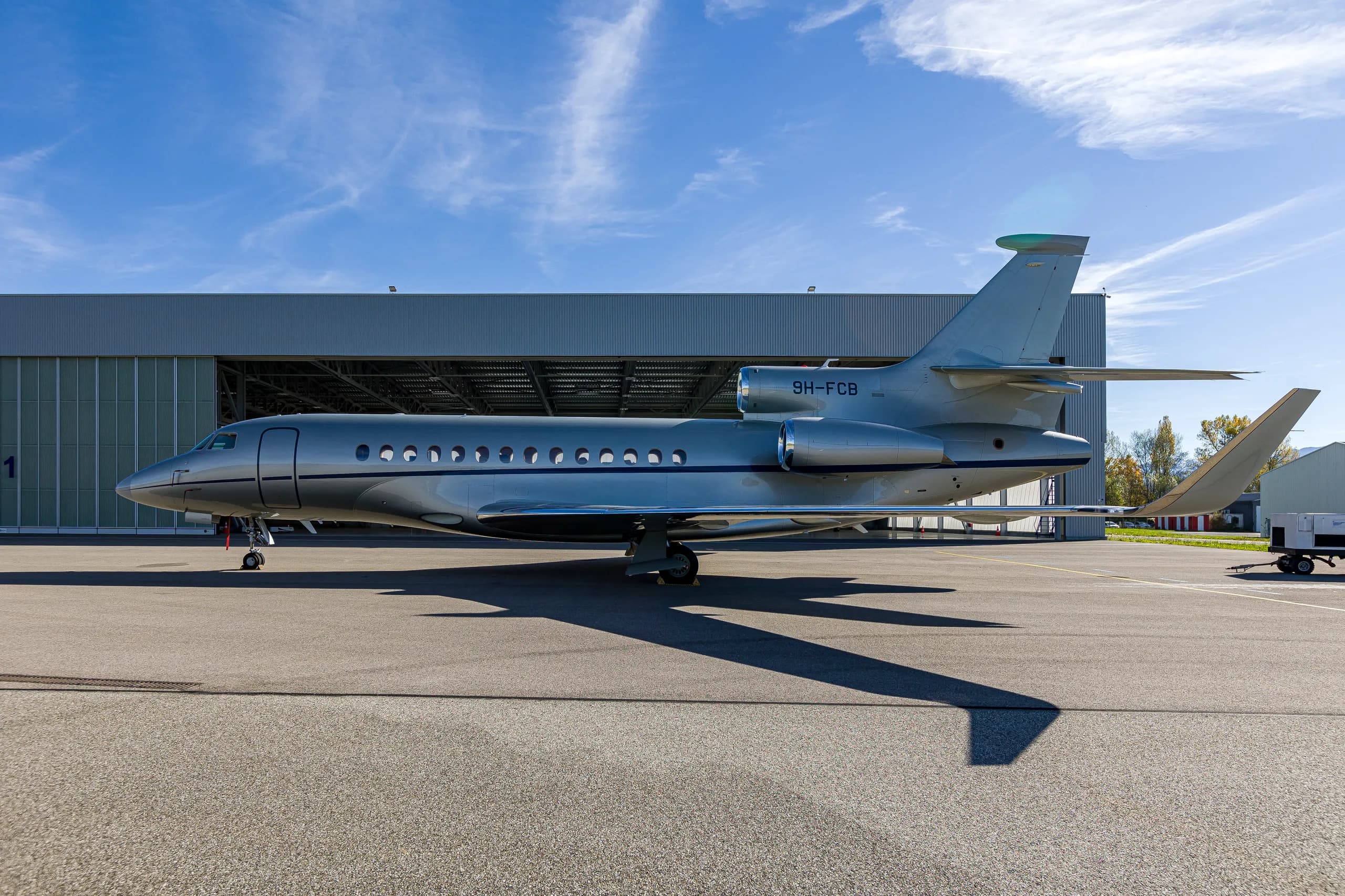 A silver Dassault Falcon 900EX business jet with blue stripes parked on an airfield next to a hangar.