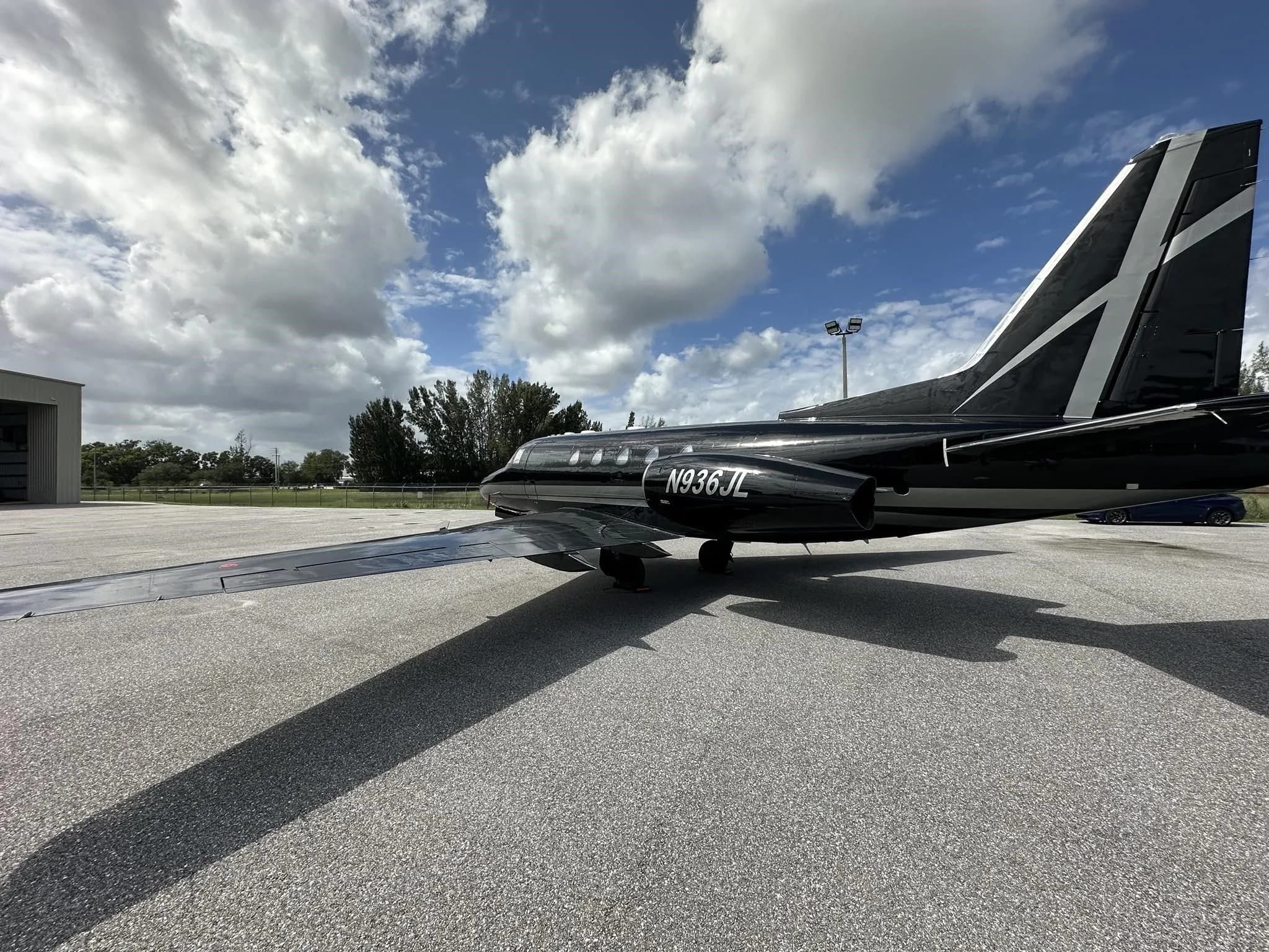 A rear-side view of a black and silver business jet, N936JL, showing its tail and engine, parked on an asphalt tarmac.