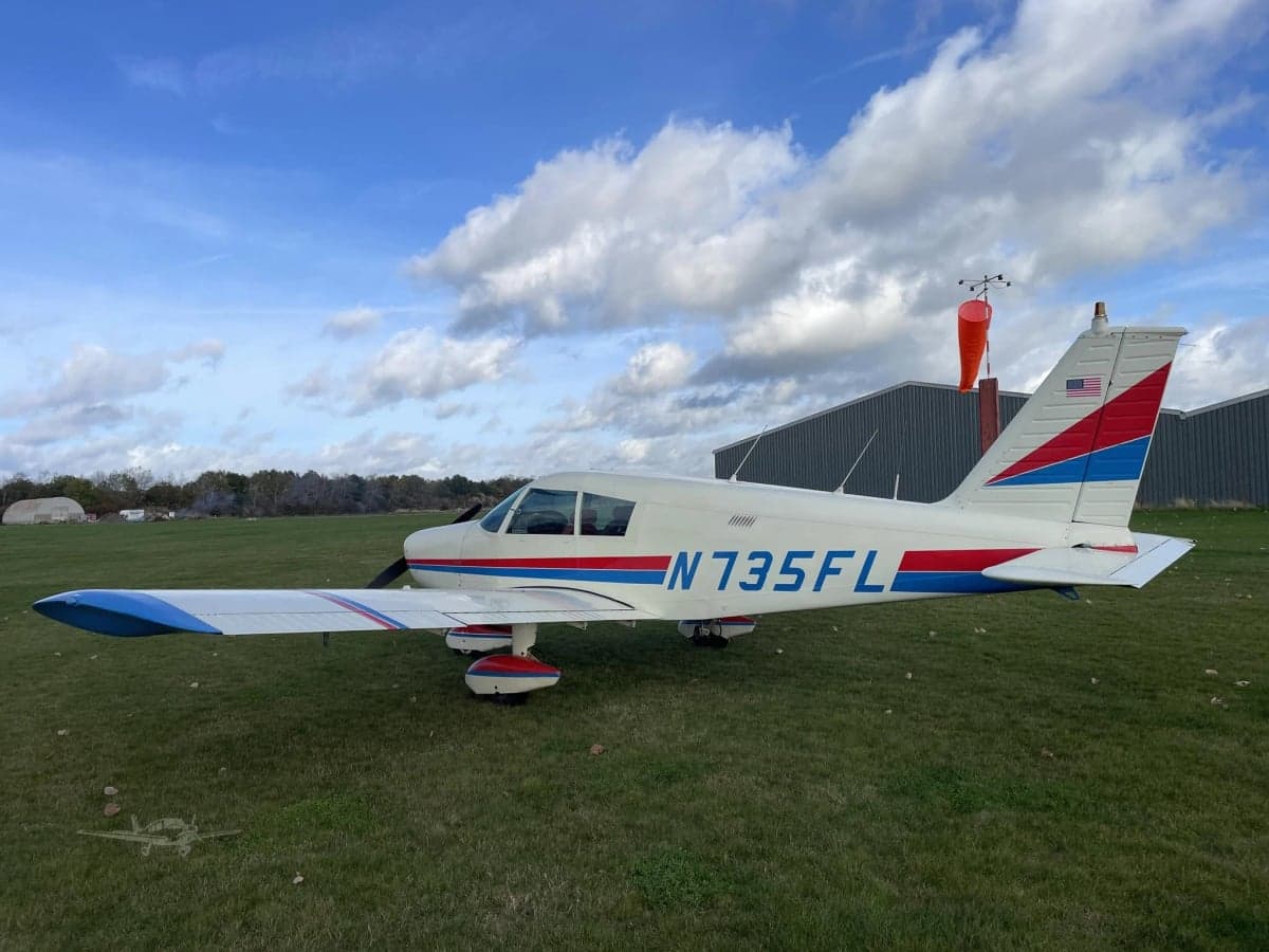 A Piper PA-28-181 Archer II aircraft, registration N735FL, is parked on a grassy field with a windsock nearby.