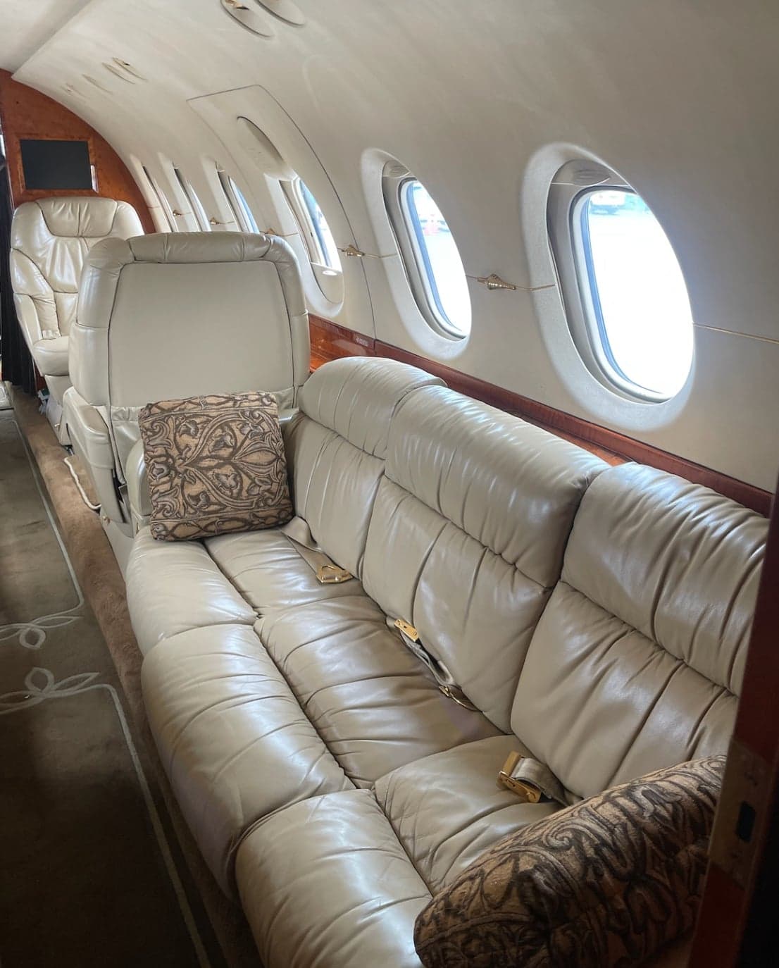 The interior cabin of a business jet, showing a long beige leather sofa with patterned pillows and several windows.