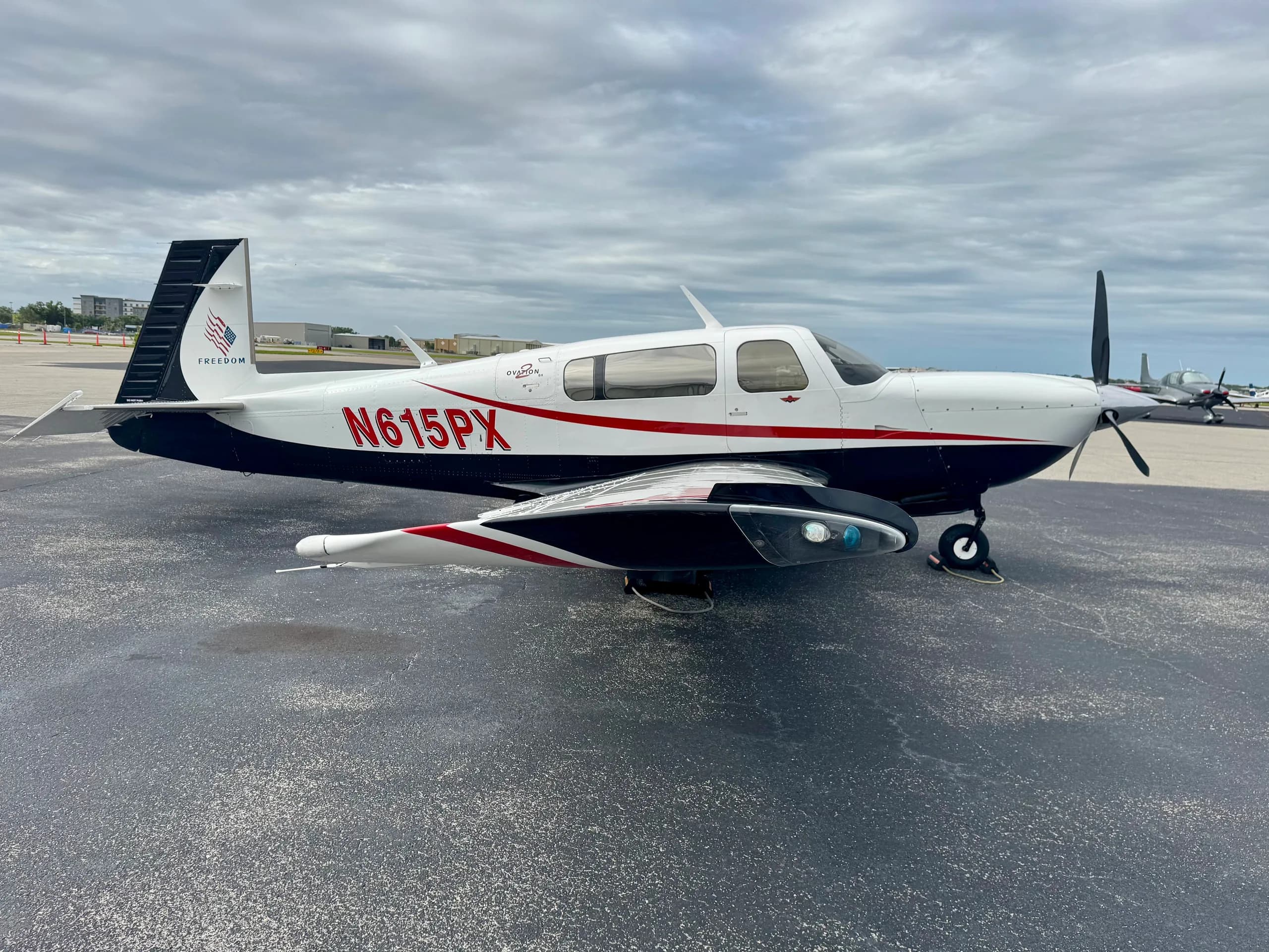 A white and black Mooney Ovation GX aircraft with red N-number N615PX parked on an asphalt ramp under a cloudy sky.
