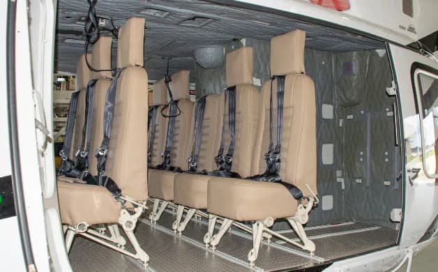 The interior of a helicopter cabin shows two rows of tan passenger seats with black harnesses.