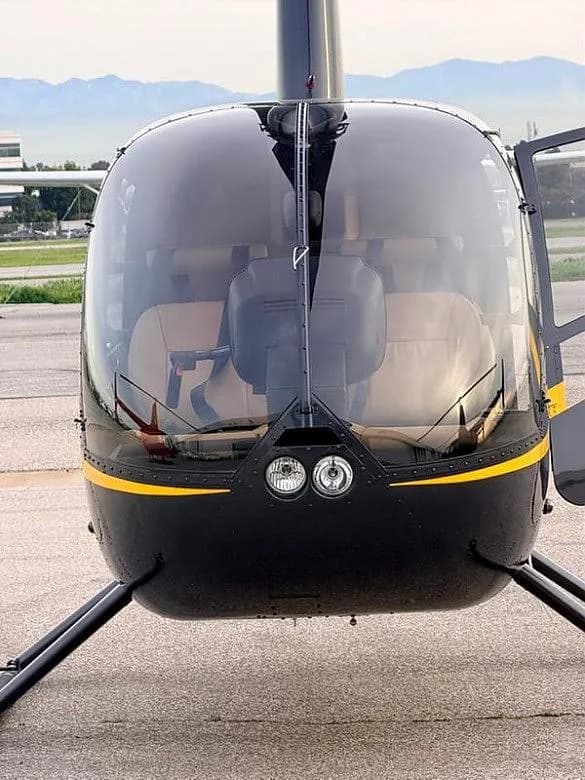A front view of a black Robinson R44 helicopter with a yellow stripe, showing the cockpit and tan seats.