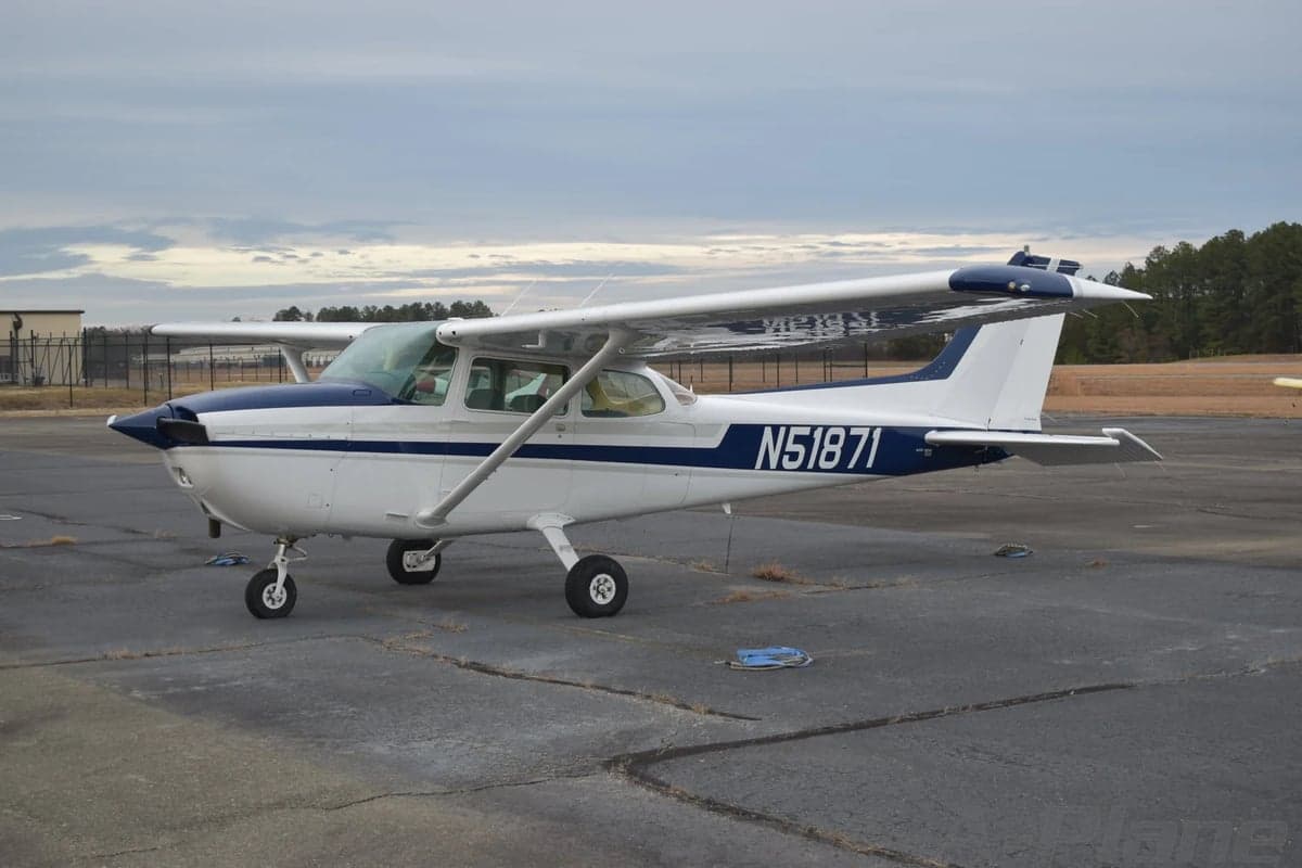A white Cessna 172 with blue and grey stripes is parked on a tarmac, viewed from the front-left.