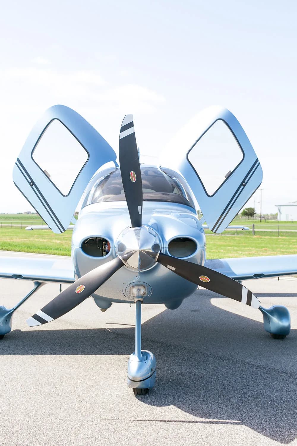 A front view of the light blue Cirrus SR22 aircraft with its gull-wing doors open on an airfield.
