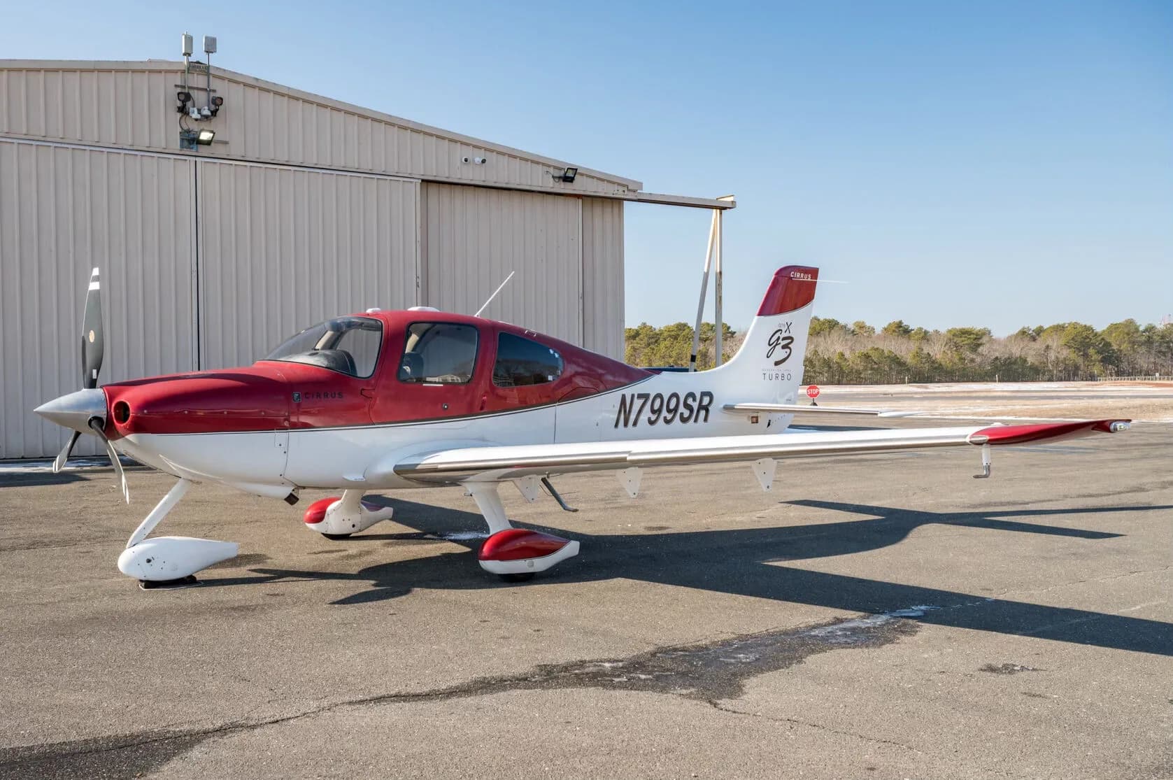 A red and white Cirrus SR22T aircraft, N799SR, parked on an tarmac next to a hangar.