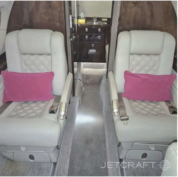 Two cream-colored cabin seats with pink pillows face a dark wood galley or lavatory area.
