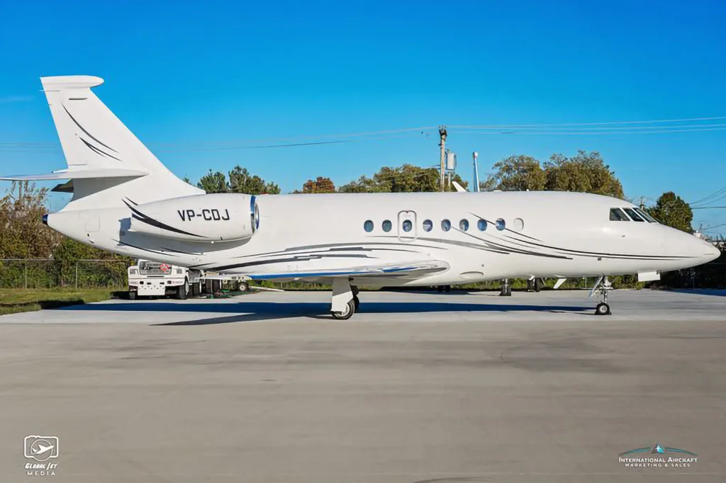 A white Dassault Falcon business jet with black and silver livery, registration VP-CDJ, parked on an tarmac with a ground vehicle behind it.