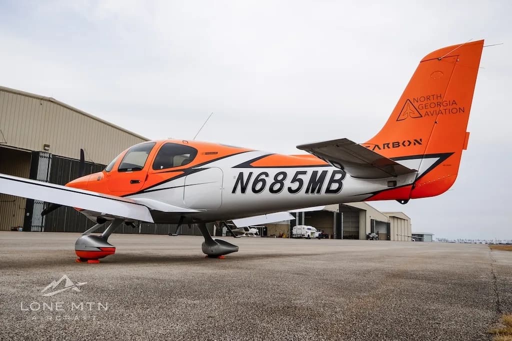 A side view of the orange and silver Cirrus SR20 aircraft N685MB parked near a hangar.
