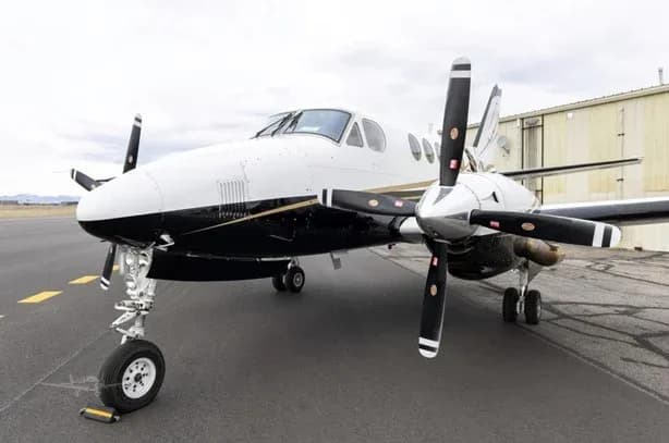 A white and black twin-turboprop aircraft, likely a King Air, is parked on an asphalt tarmac with a hangar in the background.