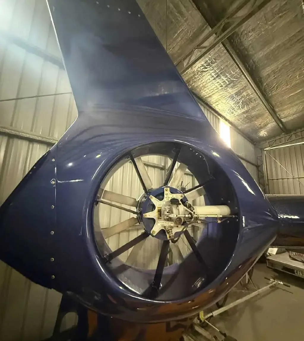 The blue tail section of a helicopter, featuring its shrouded fenestron tail rotor, inside a hangar.