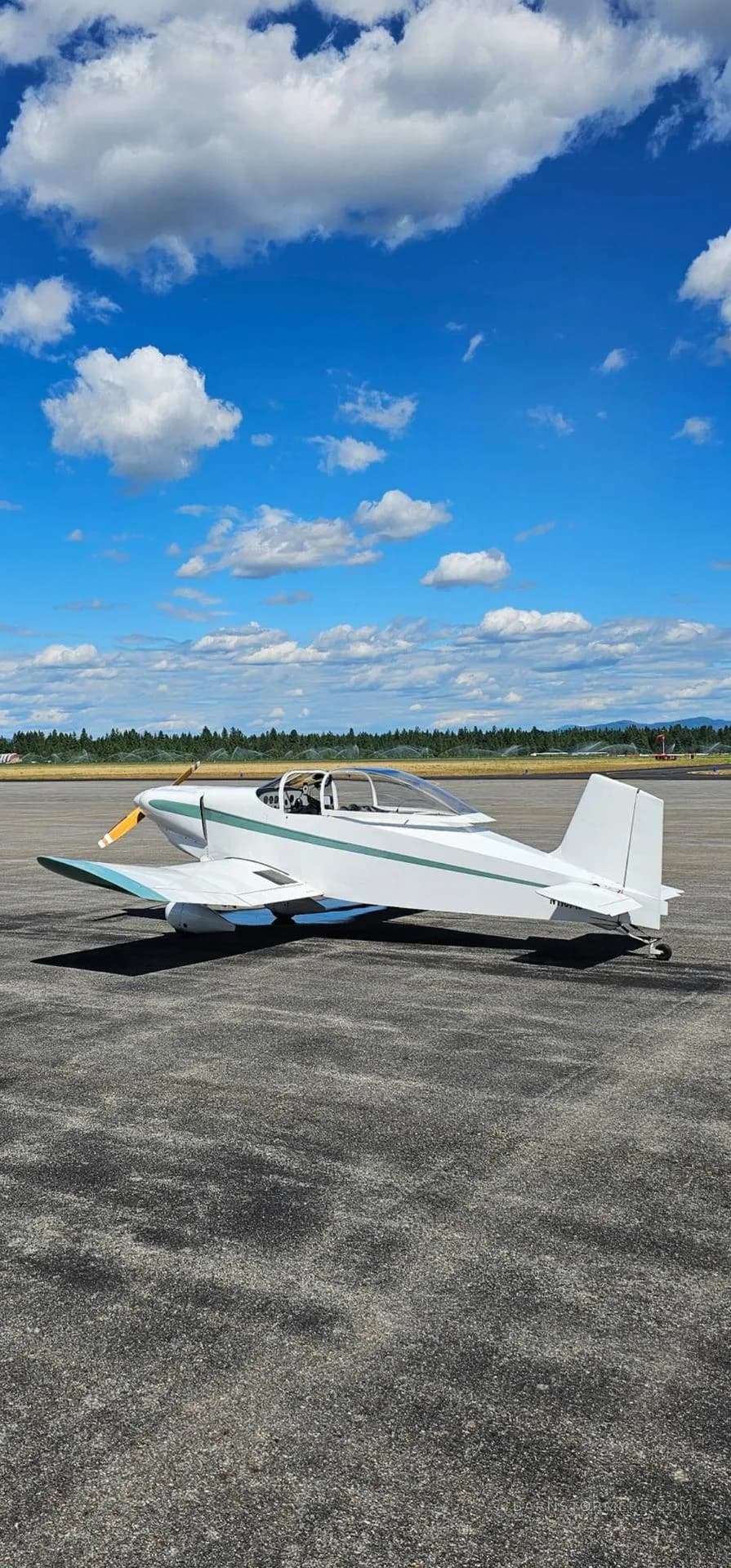 A white single-engine aircraft with teal stripes is parked on an asphalt runway under a blue sky with scattered clouds.