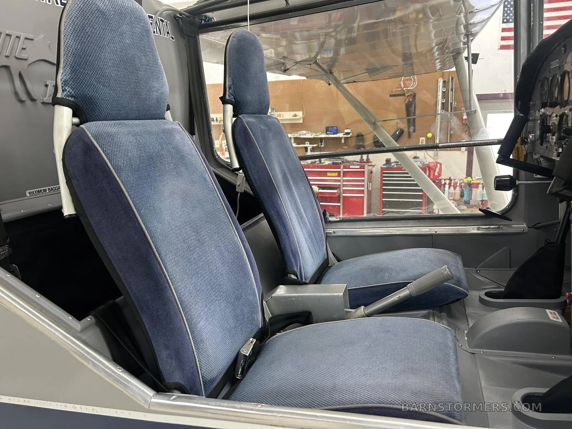 Two blue fabric aircraft seats are visible in the cabin, with a view into a hangar workshop.