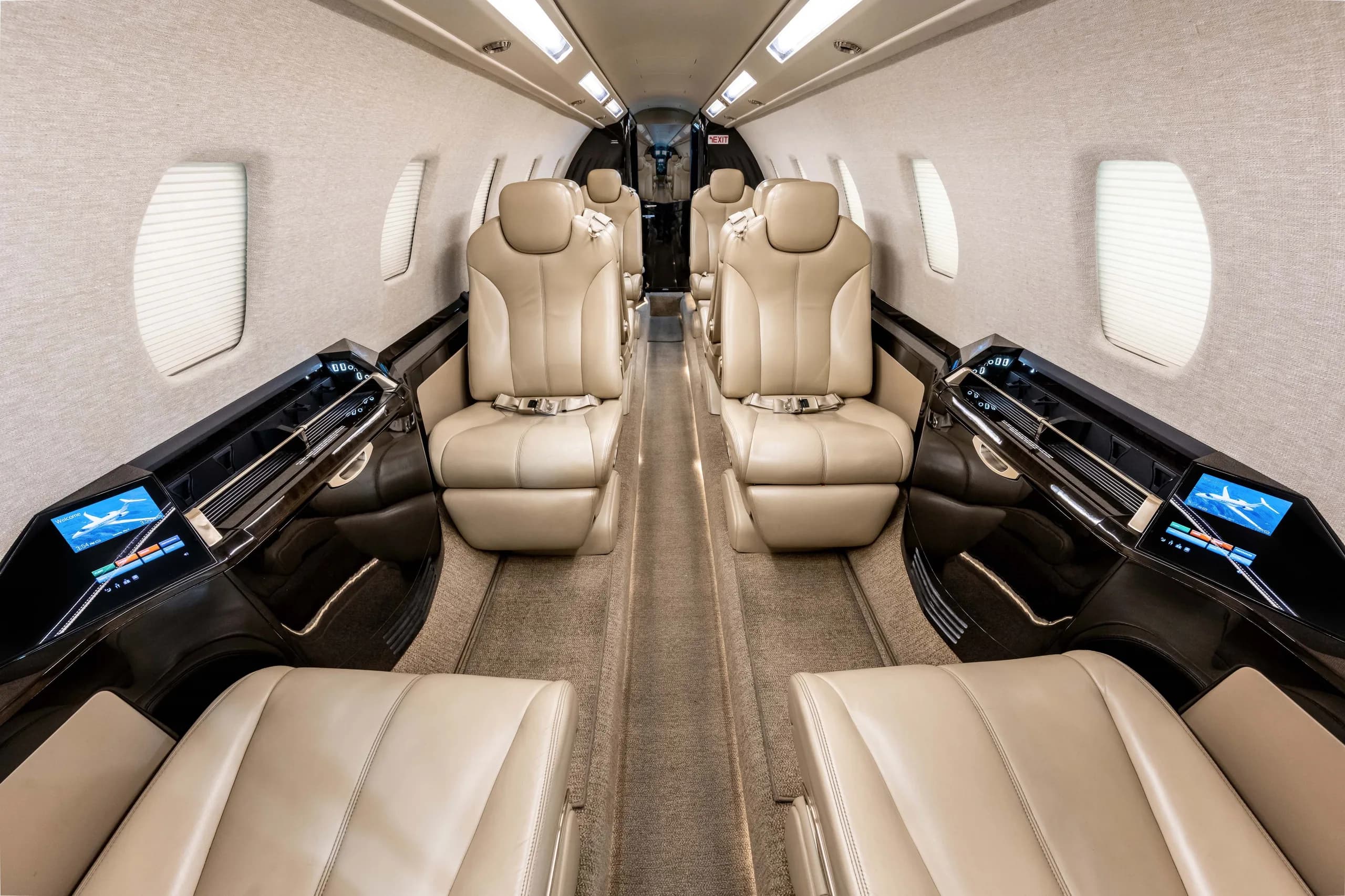 The interior cabin of a business jet with light tan leather seats arranged in a club configuration, dark wood accents, and individual display screens.