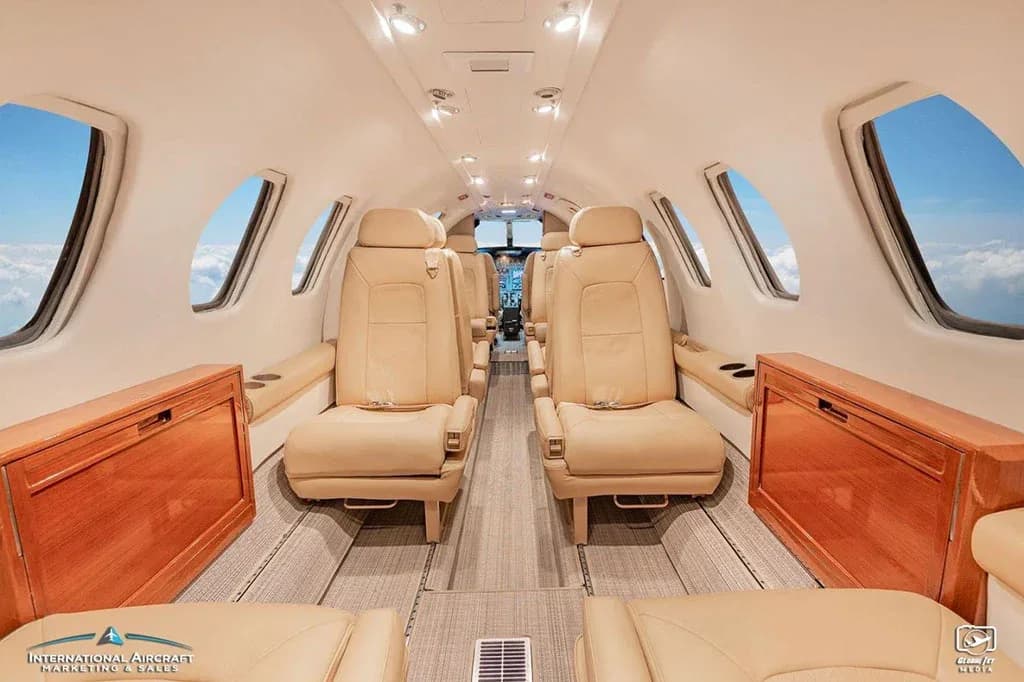 A wide-angle view of the business jet cabin with beige leather seating and wooden cabinetry.