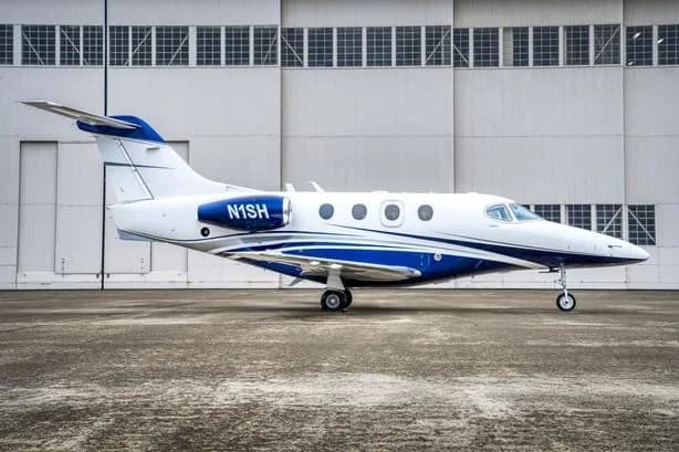 A white and blue business jet, tail number N1SH, parked on the tarmac in front of a large hangar.