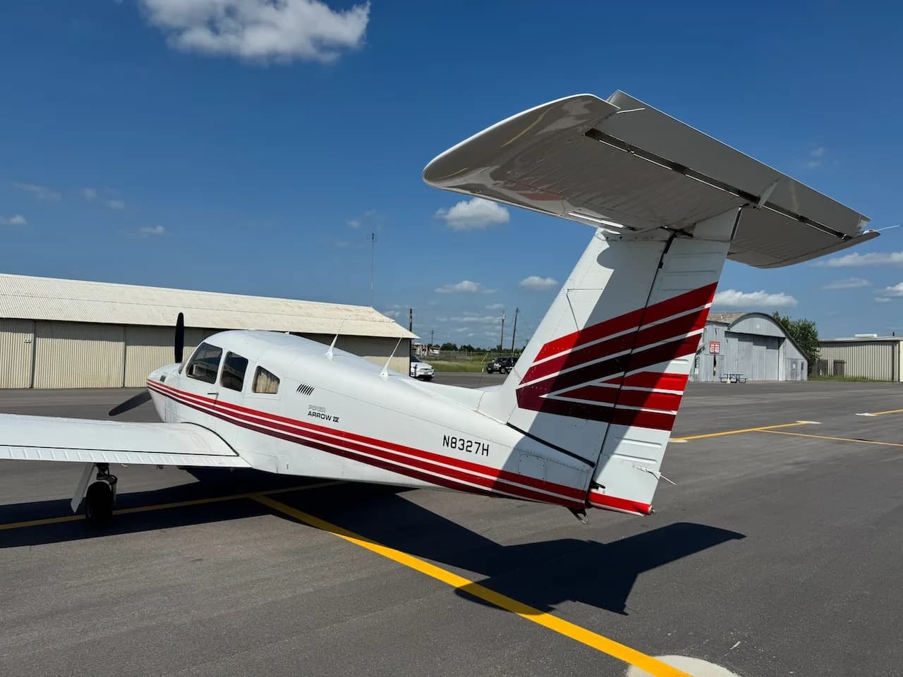 A white Piper Arrow II aircraft with red and brown stripes parked on an asphalt tarmac, viewed from the rear left.