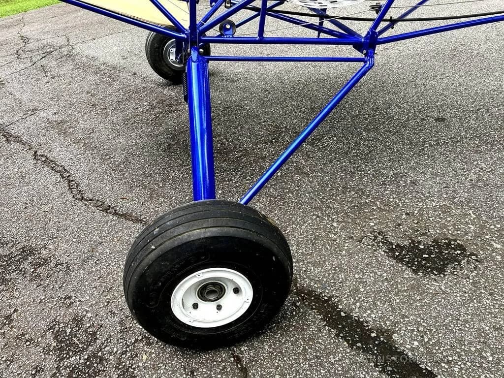 A close-up of the blue aircraft frame's landing gear with a large black tire on a paved surface.