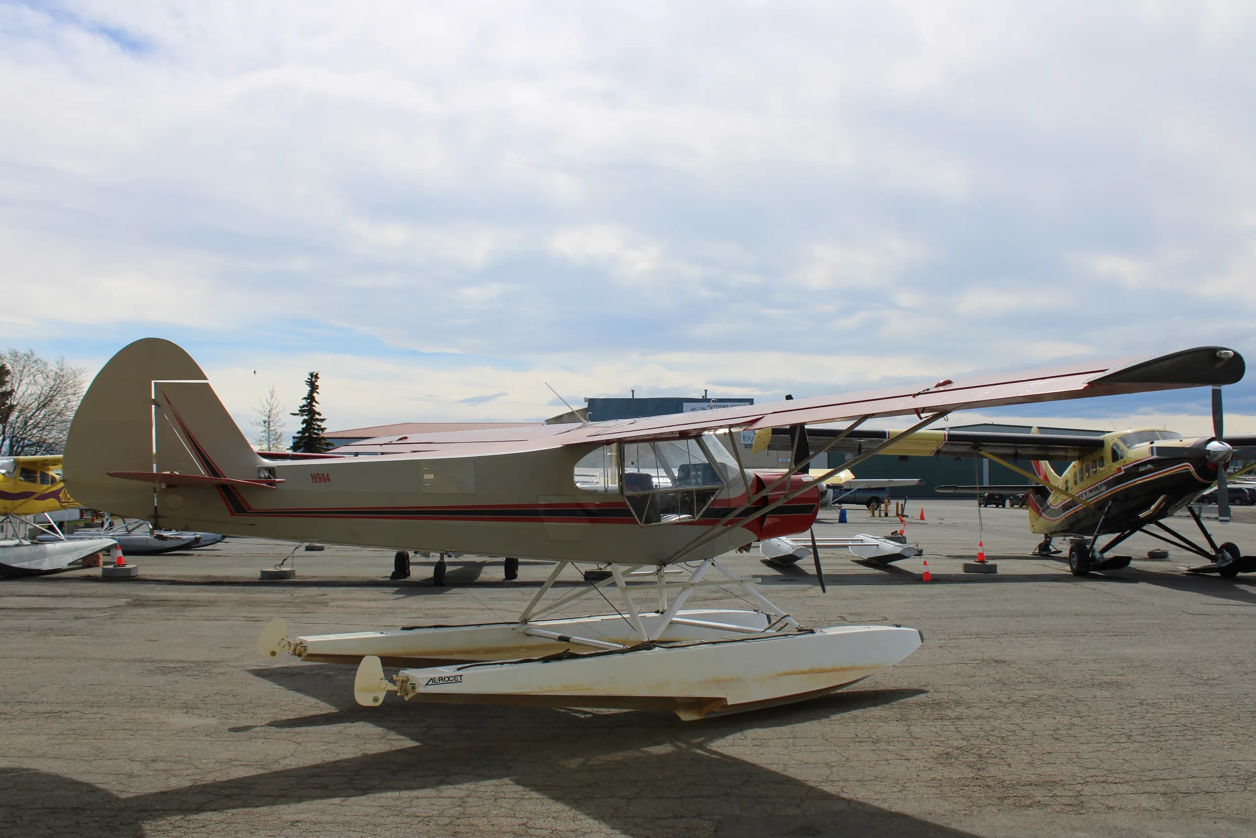 A red and silver float plane is parked on the tarmac, side view with other aircraft.