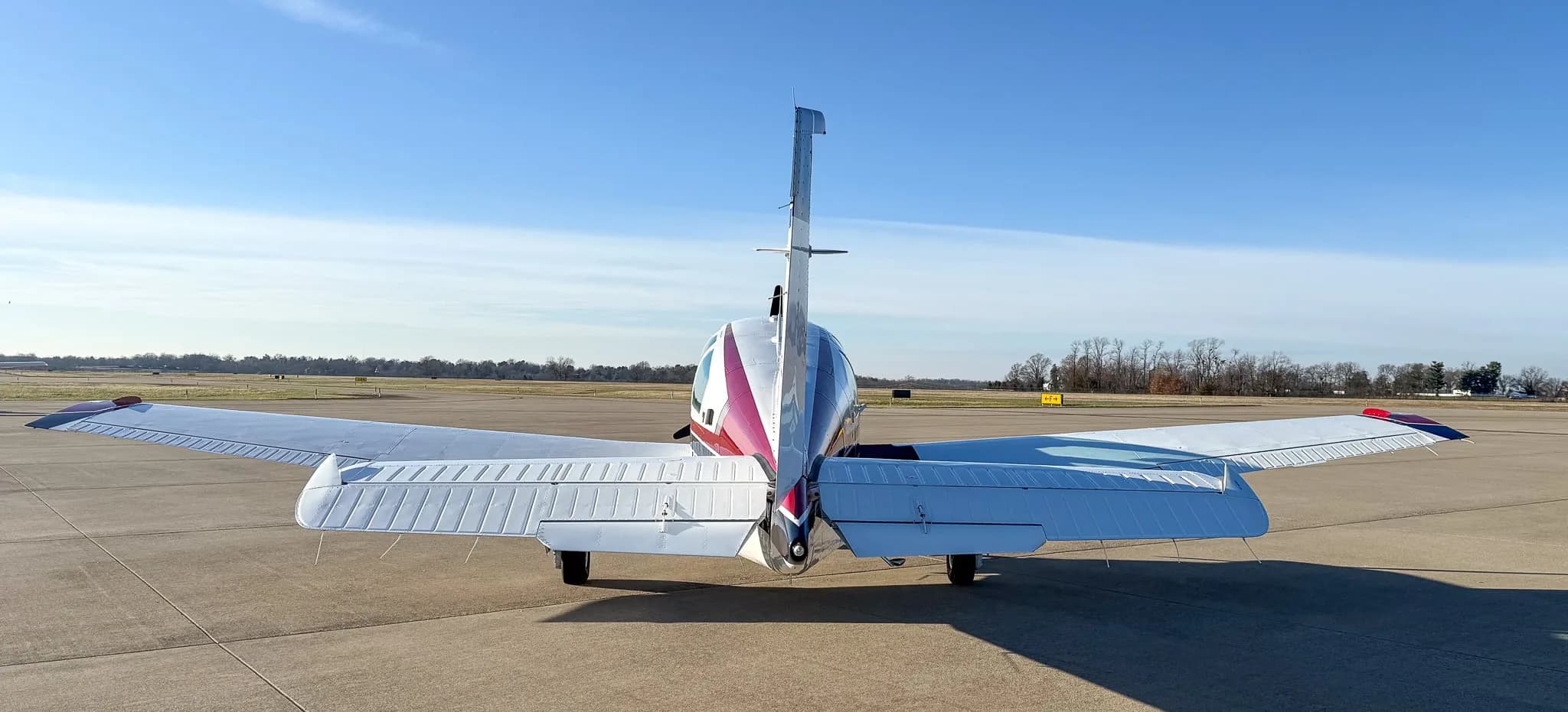 Rear view of a white Beechcraft Bonanza with red and blue tail stripes parked on an airfield.