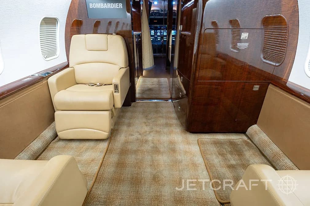 The aircraft cabin looking forward, showing a beige leather seat and dark wood cabinetry.