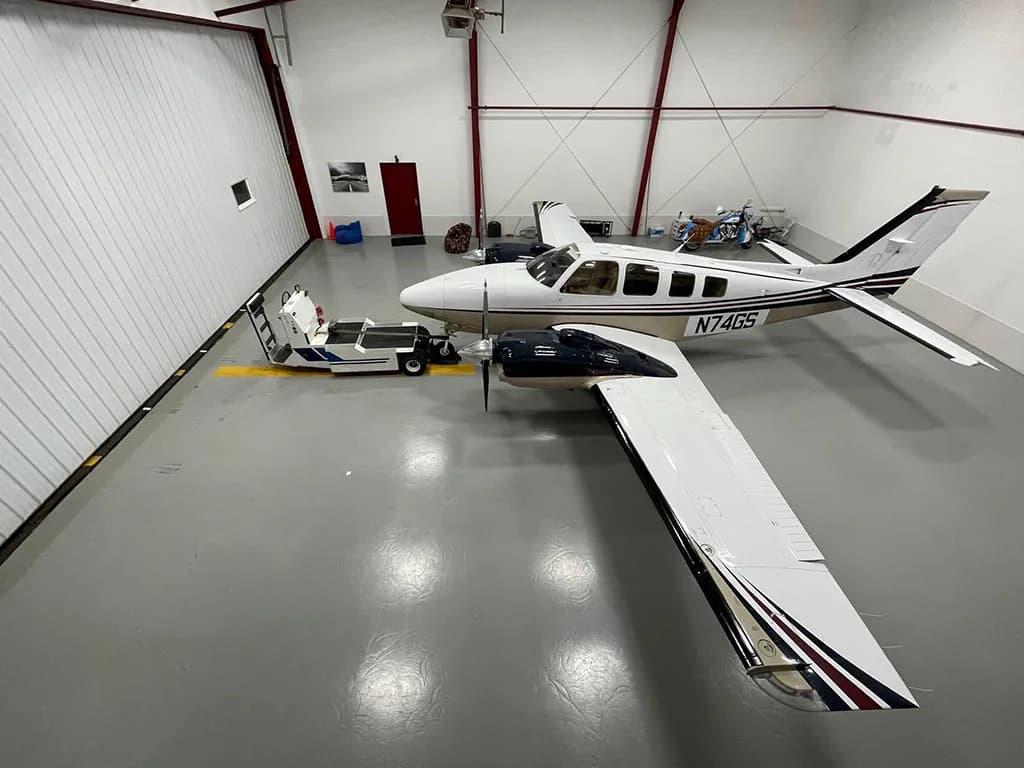 An overhead view of a white and tan twin-engine aircraft with dark blue accents parked in a hangar next to a tow vehicle.