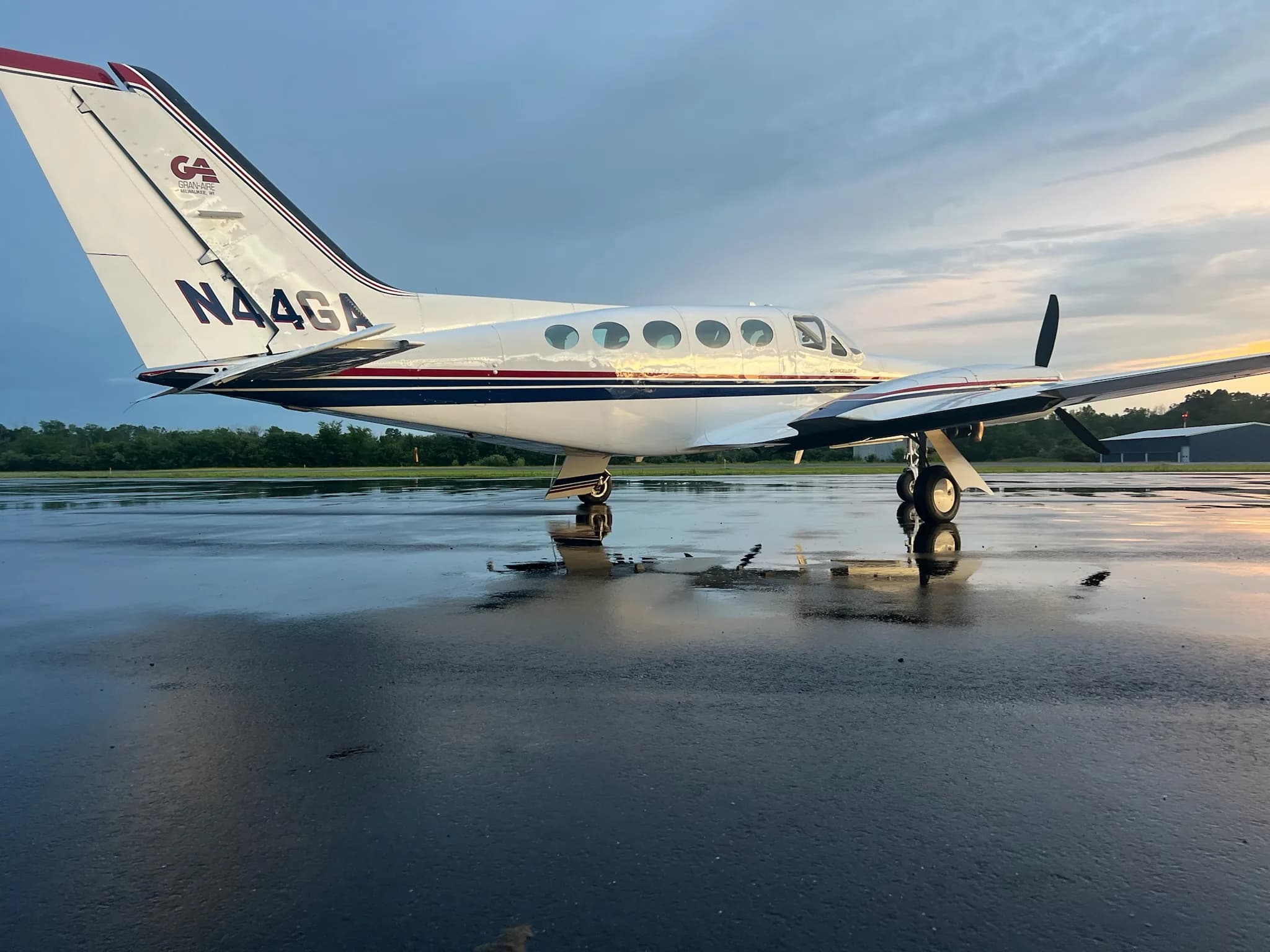 A white Cessna 414 Chancellor III with dark blue and red stripes parked on a wet tarmac at sunset.