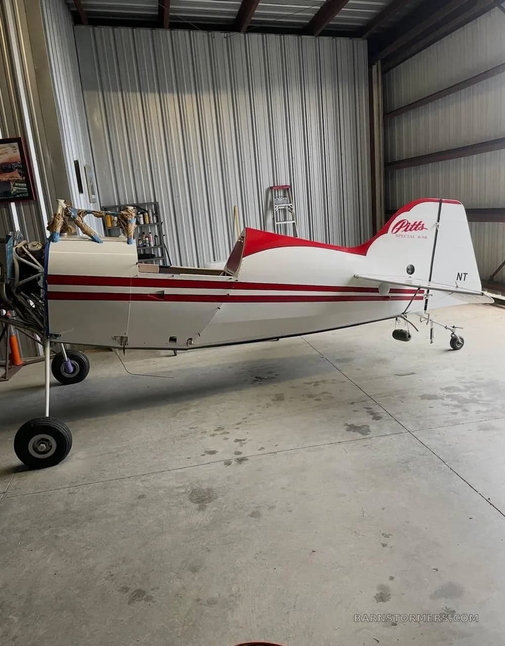 A side view of a white and red Pitts Special fuselage, with wings removed, parked in a hangar.