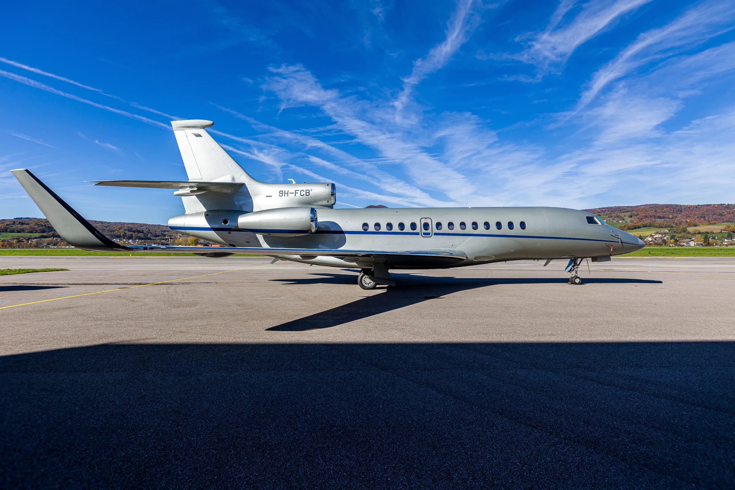 A silver Dassault Falcon 900EX business jet with blue stripes parked on an airfield, casting a long shadow.
