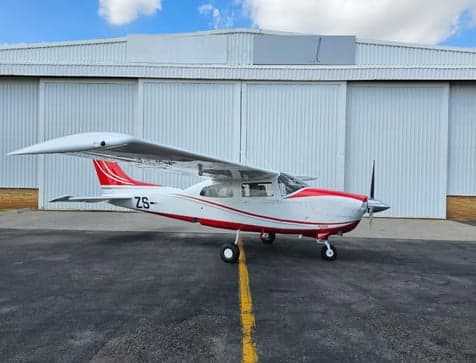A white and red Cessna 210 Centurion aircraft with ZS- registration is parked on an airfield in front of a white hangar.
