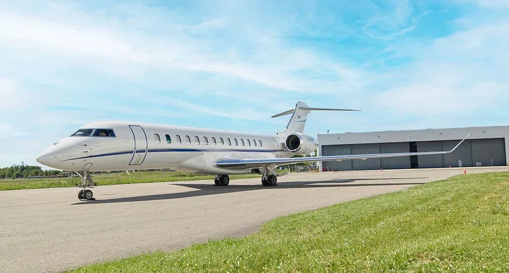 A white Bombardier Global business jet with blue stripes on the tarmac next to a hangar under a blue sky.