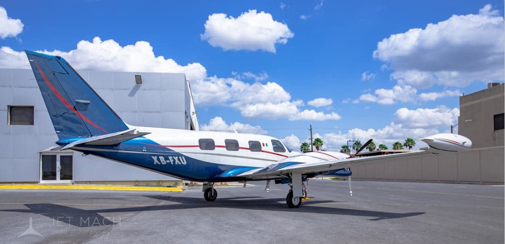 A white and blue twin-engine propeller aircraft, registration XB-FXU, is parked on a tarmac next to a building.