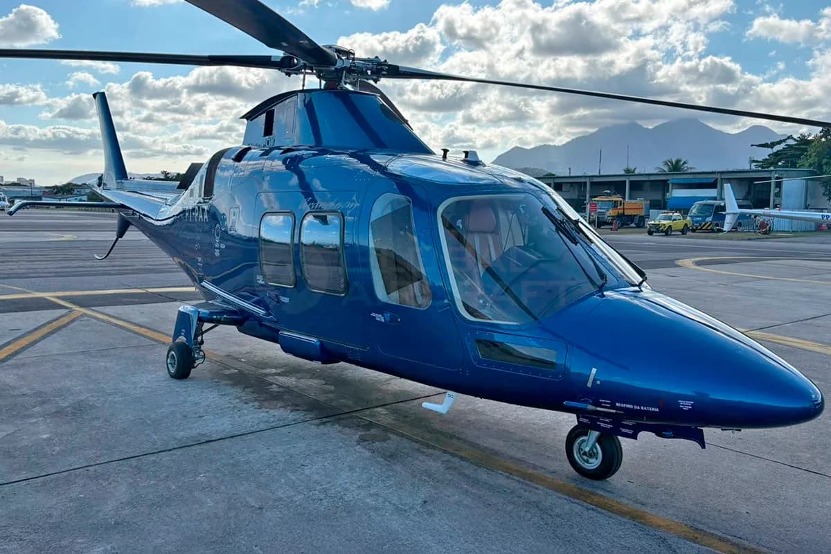 A front-side view of a blue Leonardo AW109 helicopter (PT-PAA) parked on an airfield under a cloudy sky.