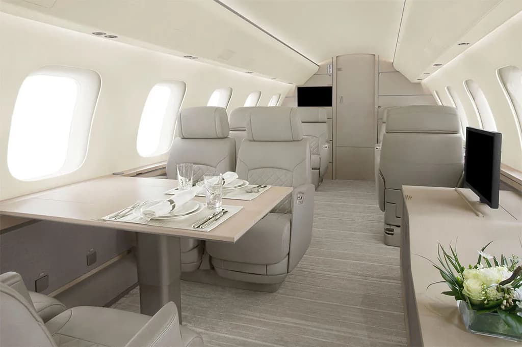 A spacious aircraft cabin with a dining table set for two, surrounded by light grey leather seats and a floral arrangement.