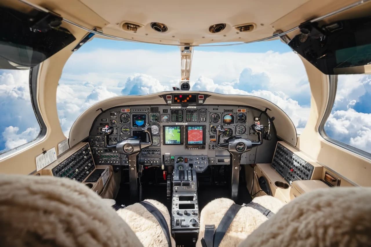 The cockpit of a business jet, showing the instrument panel with multiple displays and controls, with clouds visible through the windshield.