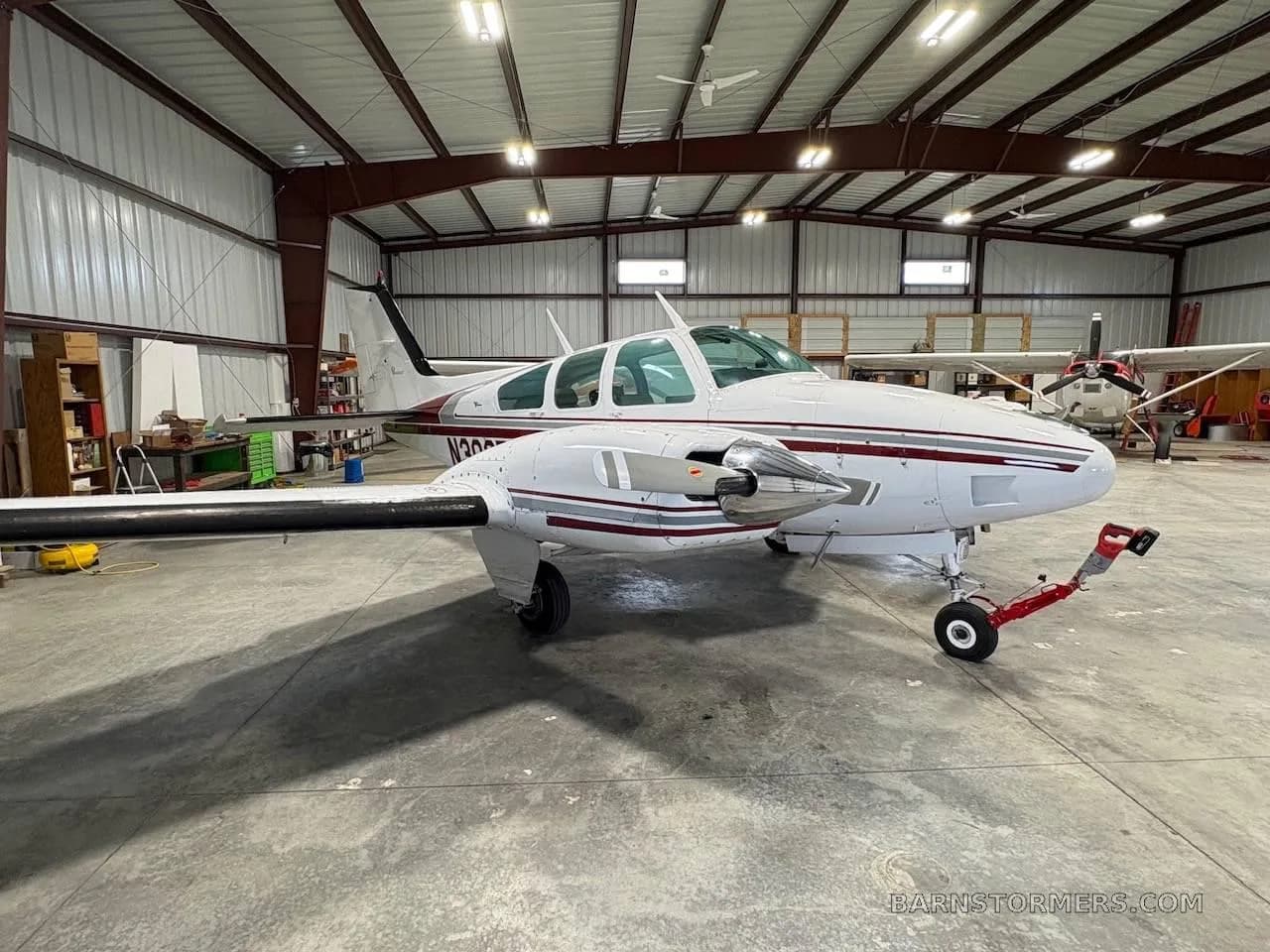 A white twin-engine propeller aircraft with red and grey stripes, seen from the front-right in a hangar.