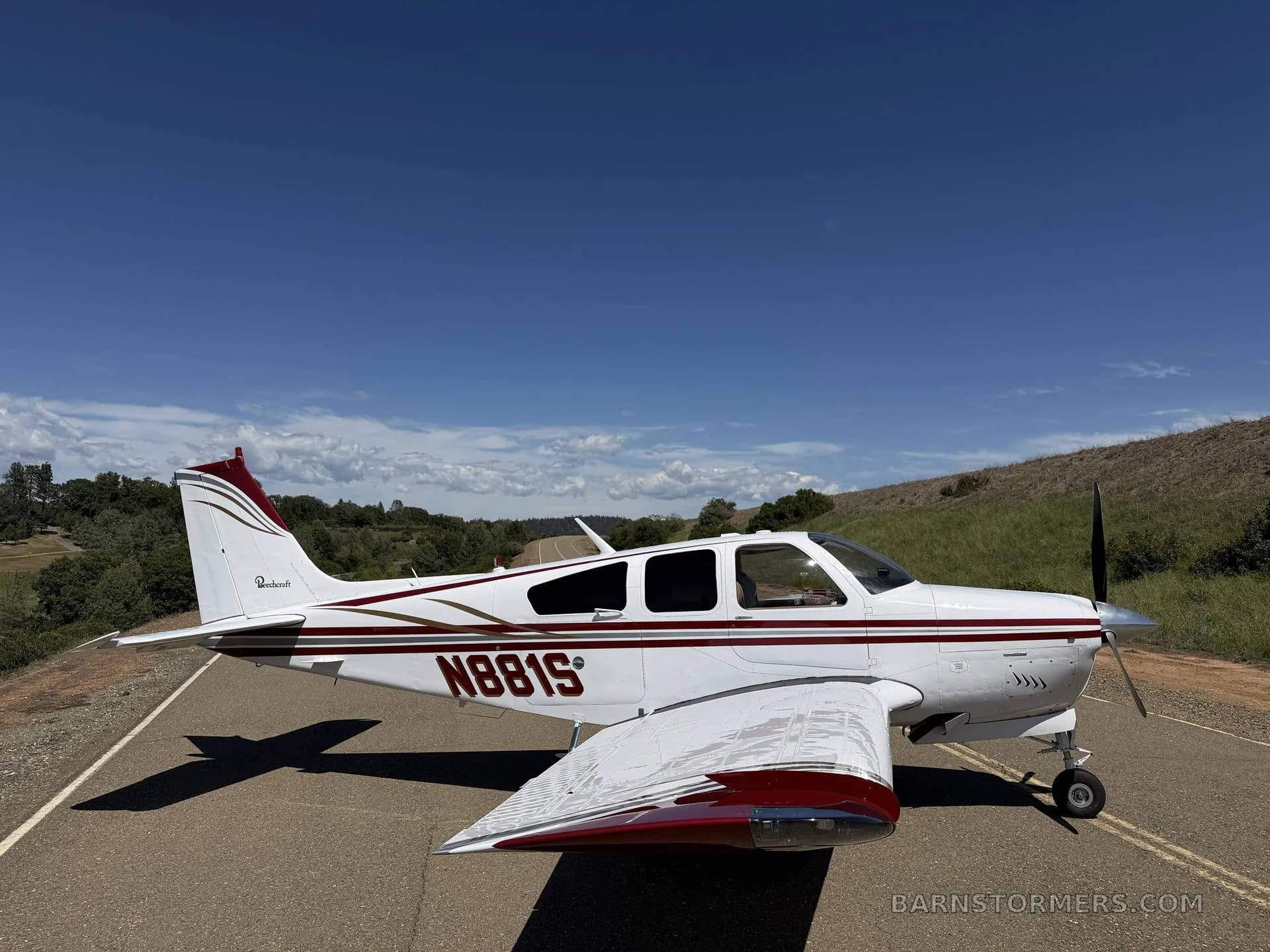 A white Beechcraft Bonanza N881S with red and gold stripes parked on an asphalt surface under a clear blue sky.