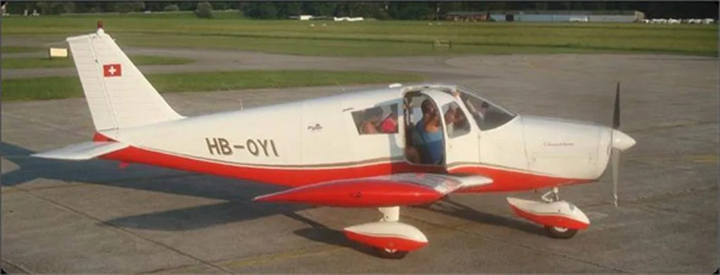 A white and red Piper Cherokee aircraft (HB-OYI) with its door open, parked on an airfield with people inside.