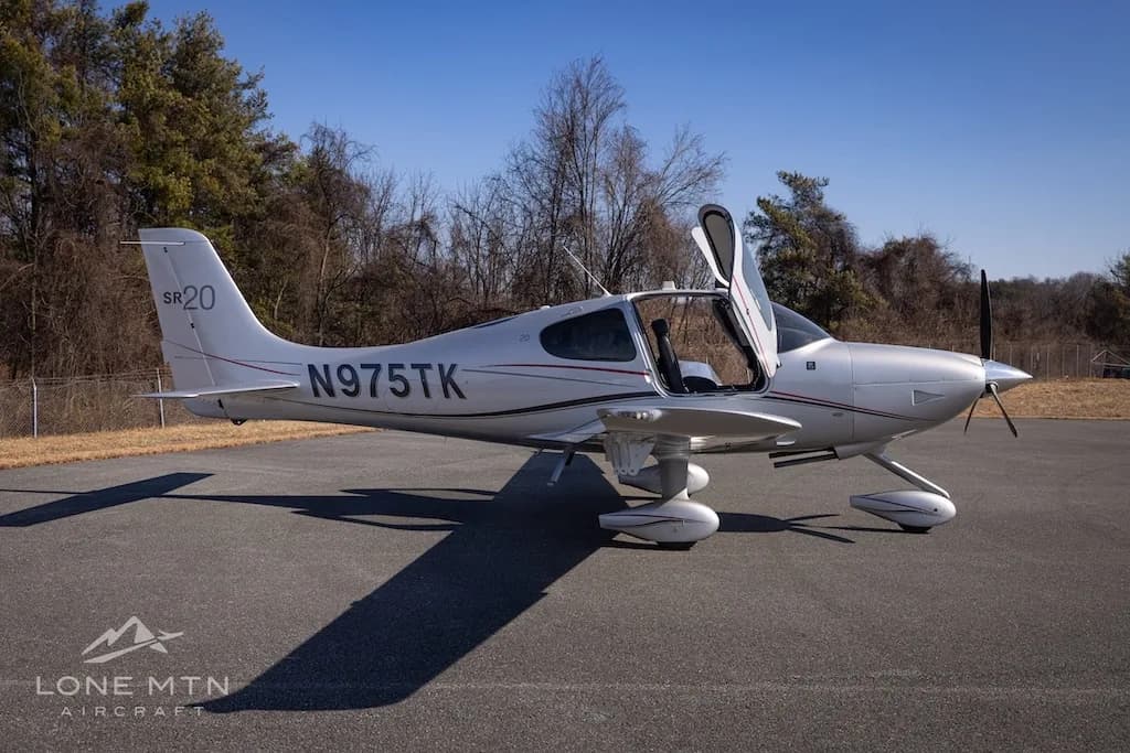 A silver Cirrus SR20 aircraft with registration N975TK parked on an asphalt runway, viewed from the right side with doors open.