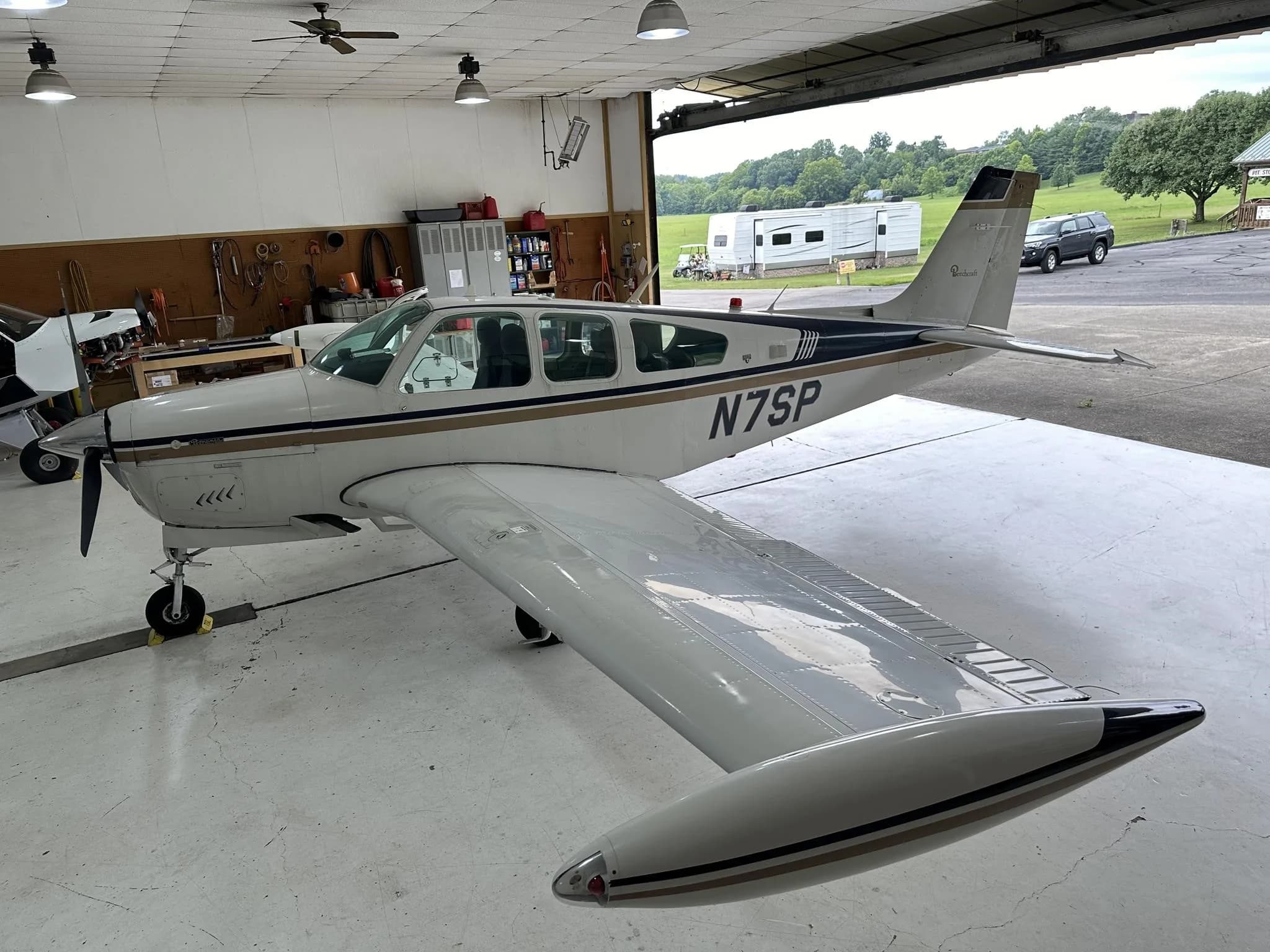 A white single-engine propeller aircraft with gold and black stripes, tail number N7SP, is parked inside a hangar with the door open to a grassy field.