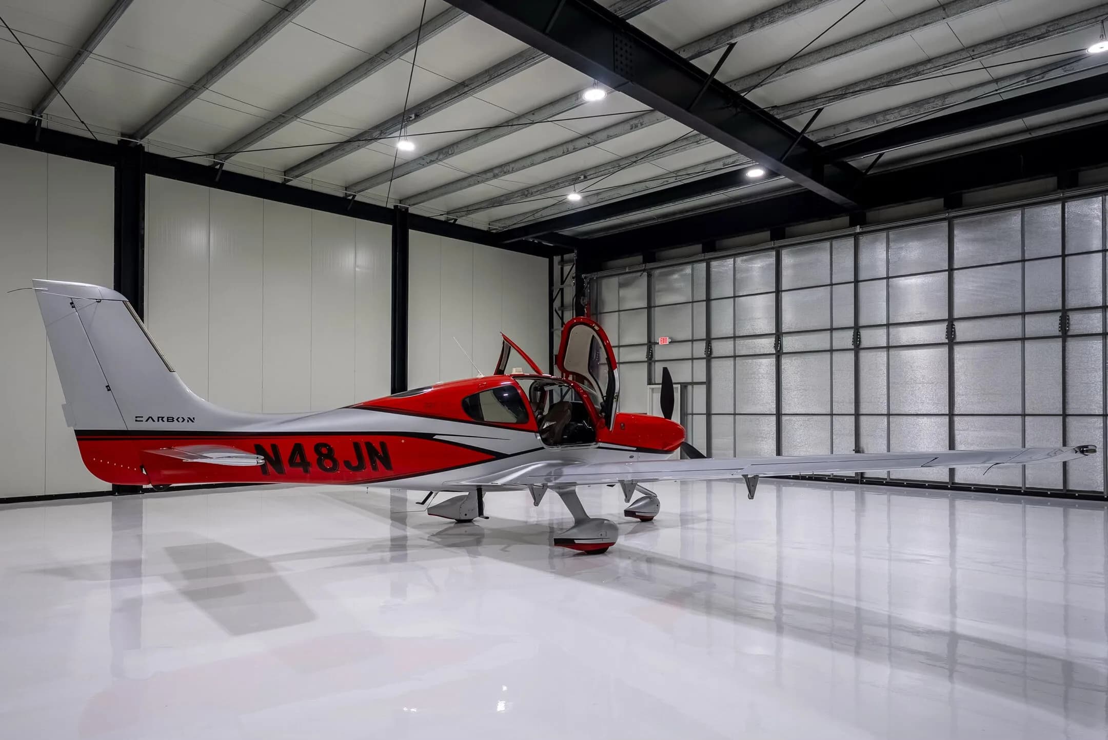 A red and silver Cirrus SR22T Carbon aircraft with registration N48JN and open doors, parked in a hangar with a large door in the background.