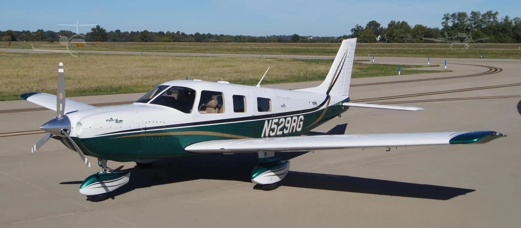 A white and green Piper 6XT aircraft (N529RG) with gold accents is parked on an airfield under a clear blue sky.